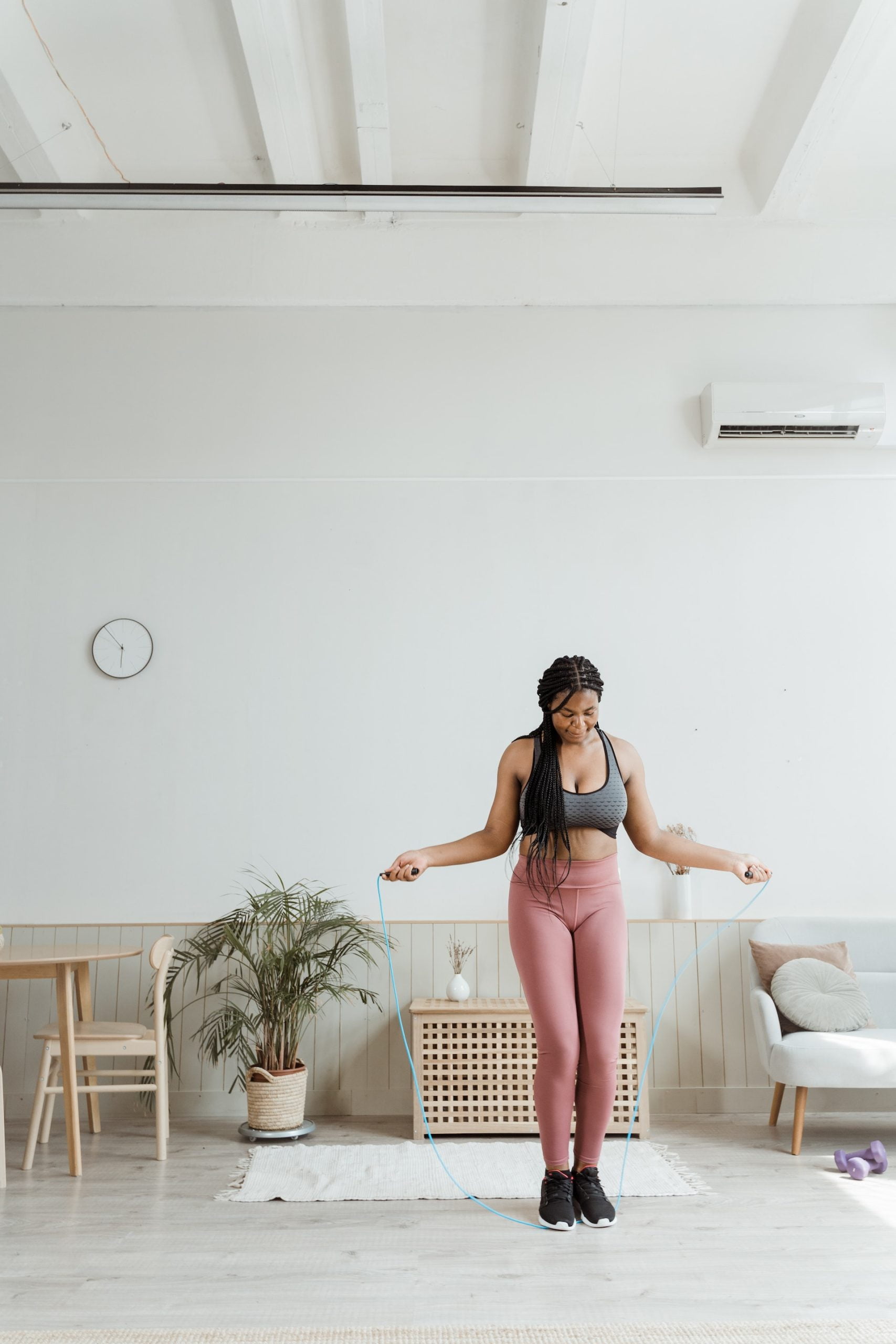 A woman jumping rope in a bright room, wearing a sports bra and leggings, with a plant and furniture in the background.