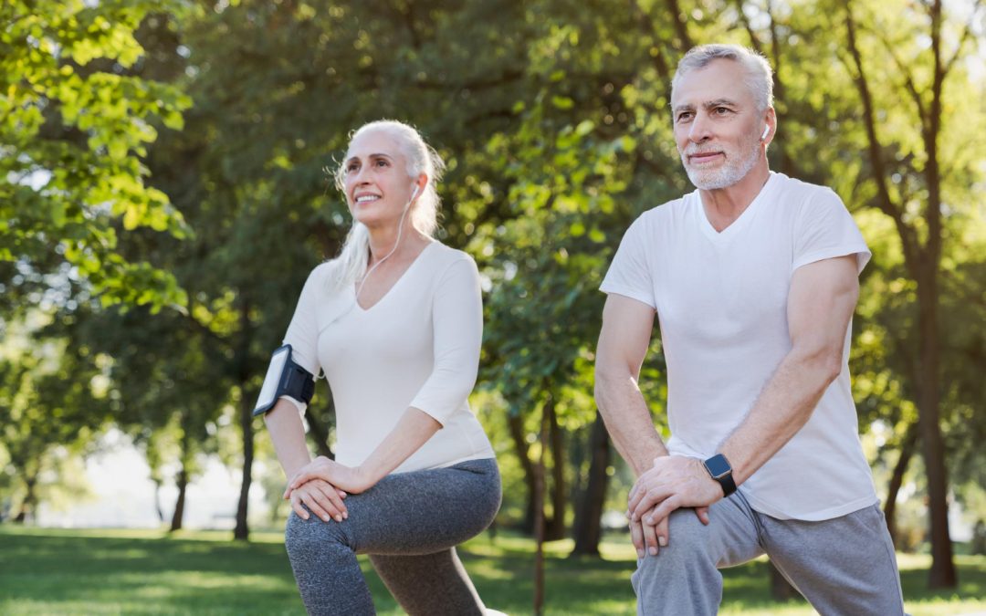 Two seniors doing lunges in a park, wearing workout clothes and earphones, with trees in the background.