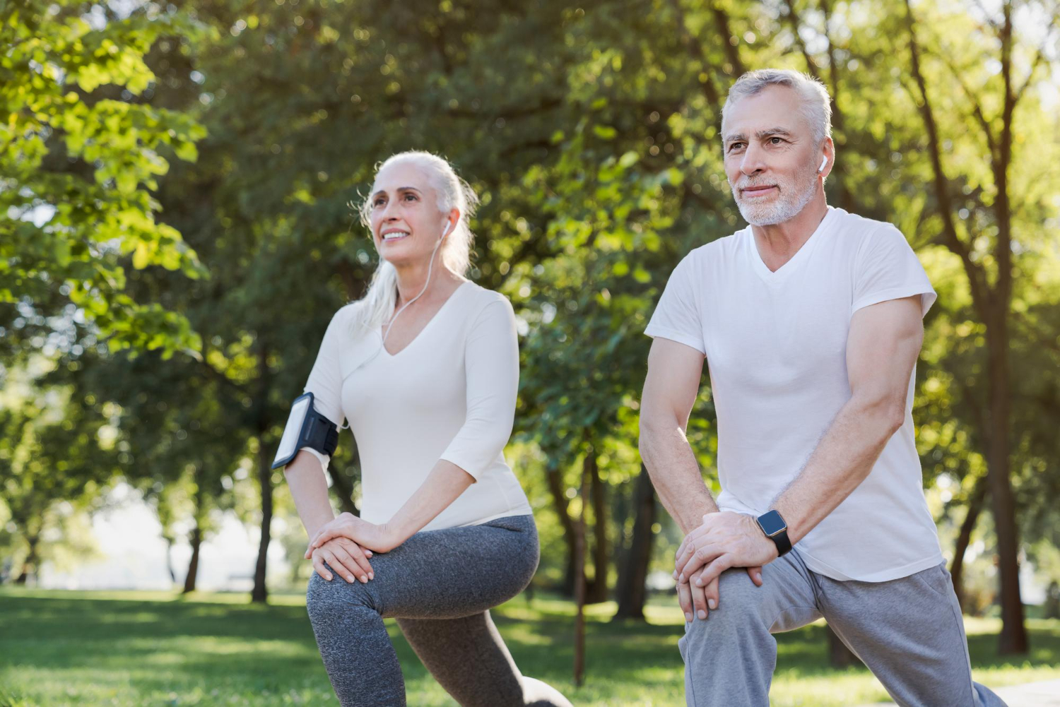 Two seniors doing lunges in a park, wearing workout clothes and earphones, with trees in the background.