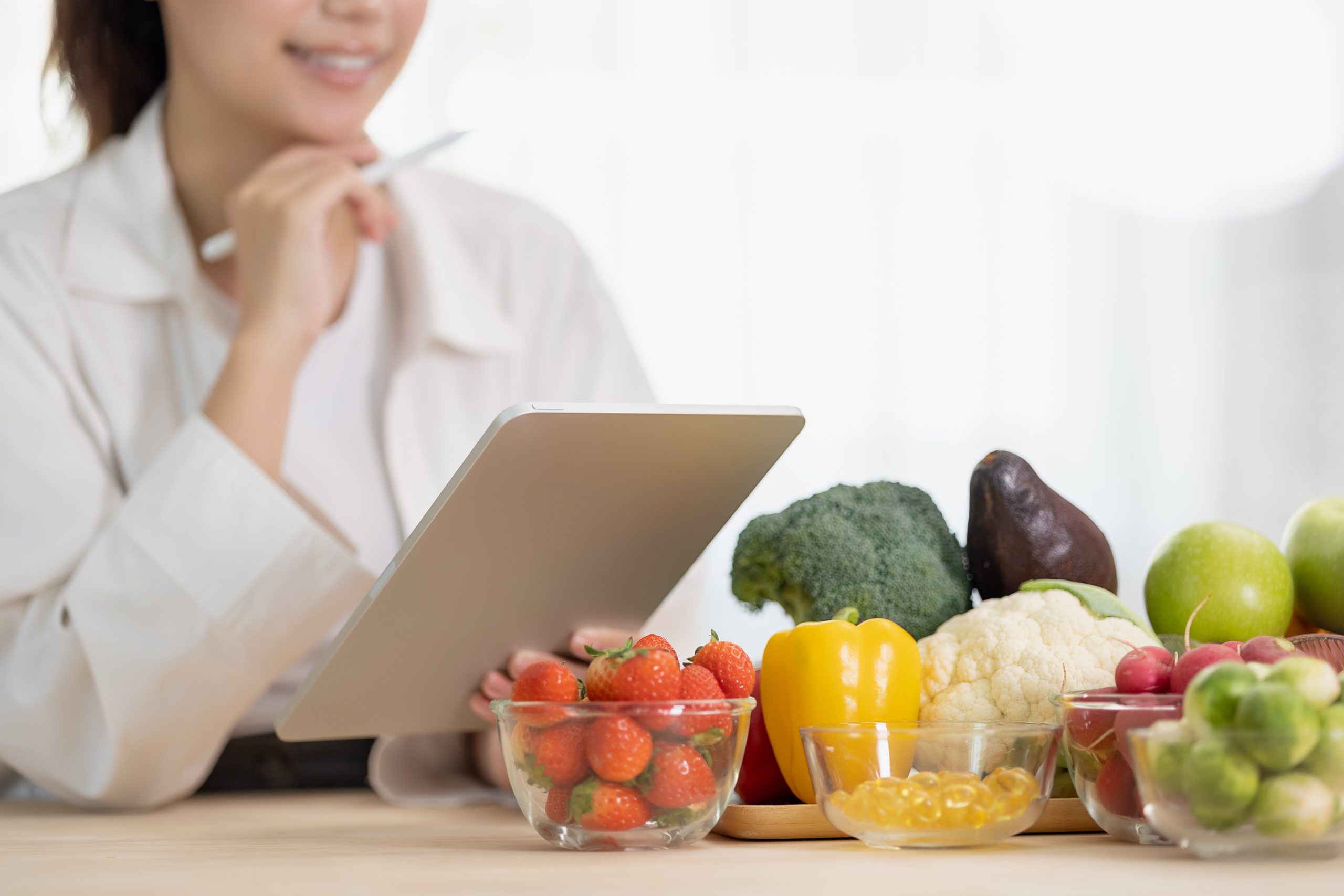 A person holding a tablet near a table with assorted fruits and vegetables, including strawberries and broccoli. Nutrition Counselor